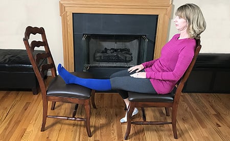 Photo of a woman sitting in a chair correctly with her left leg elevated and resting on a second chair